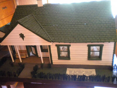 A replica of a white building with a green roof. A tag on the building reads "buil in 1939 by the C.C.C. for Bob and Madge Corrigan, children of their supervisor. This is a replica of a typical [illegible].