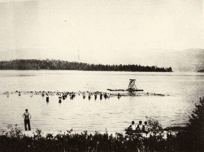 View of CCC men swimming in Kalispell Bay. There is a life guard standing watch and a row of three men sitting on the shore in the foreground. Back of photo reads: 'CCC Swimming lessons at Kalispell Bay, Headquarters Camp F-142, 1935. Boys were brought to Priest Lake from other camps for swimming lessons. Les [Tam] photo. Chuck Peterson copy.