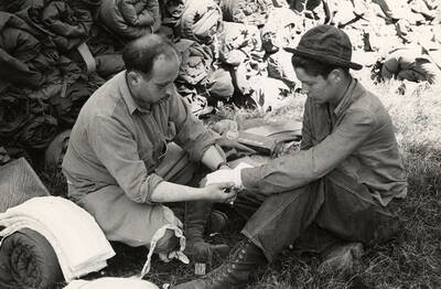 Photo of a CCC man getting his hand bandaged by another man in front of a wall of sleeping bags at the CCC Spirit Lake Fire Camp. Between the two men is a cigarette box that reads: 'Lucky Strike A Light Smoke'.