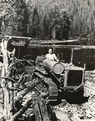 CCC man operating a cat tractor pulling a drag line to dredge sand and rock from the bottom of the Coeur d'Alene River. Sand and rock needed to make concrete for the Sissons Bridge. Back of photo reads: 'Loading gravel, Sisson's Bridge Coeur d'Alene River 1933'. Stamp on back of photo reads: 'Museum of North Idaho P.O. Box 812 Coeur d'Alene, Idaho 83814'.
