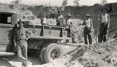 CCC work crew from Four Corners Camp pose at their worksite with shovels and a truck. Men are likely loading sand into the dump truck to be carried to a larger worksite. Writing on the photo reads: 'Company 594 Four Corners Leo's Studio'. Back of photo reads: 'Four Corners Priest River'.
