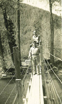 Two CCC men standing on bridge above a river. CCC Camp Big Creek #2, F-132. The bridge also holds a water line that feeds the camp. Back of the photo reads: 'water line over Uranus Creek near camp.'