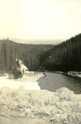 A photo of a lake surrounded by snow covered ground and forested hills.