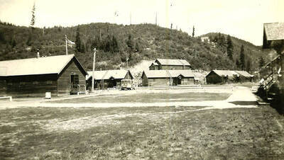 A view of the courtyard of CCC Camp Big Creek #2, F-132. There are several buildings and a flagpole around the edge of the courtyard and a truck parked on the edge. One of the buildings is an infirmary. Hills rise in the background. Back of the photo reads: 'Camp Big Creek'