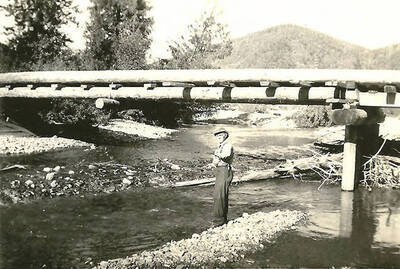 A CCC man stands under the bridge fishing in the creek. Back of the photo reads: 'Another view of bridge over Big Creek. We built them solid.'
