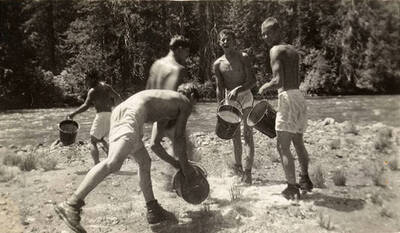 Five CCC men in white swim trunks and boots holding buckets full of water next to a river. Back of the photo reads: 'Water fight - main camp August 1938'