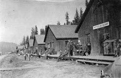 A small company of CCC enrollees lounging in front of several buildings with a connected porch. The nearest building has a sign above the door that reads: 'Matthies Restaurant' and the writing underneath the photo reads: 'Headquarters, ID'.