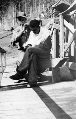 Two men in a CCC Camp. One is sitting on a bench, wearing a small smock, while the other straddles the bench and holds a razor or scissors to the first man's head. Writing below the photo reads: 'Red DeLarens O'Russel Vitalo Bear Our Company Barber'.