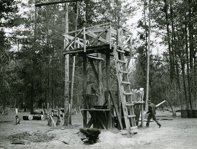 Two CCC men working with logs at a large construction (presumably a creosoting plant) in the woods. Several barrels lay in the background. Writing above the photo reads: 'CCC Enrollees preparing fence posts and telephone poles and operating creosoting plant at Stewart Flat. These products are used as far as 100 miles from this plant. By Paul S. Bieler. July 1941'