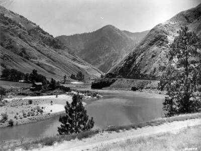 The bottom of a river valley includes a dirt road carved into the side of a hill, as well as a small farm on the other side. Hills rise into the background.