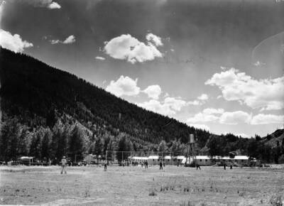 A CCC Crew plays baseball outside of a group of buildings that appears to be a CCC Camp. There is a chain link fence behind the batter, and a water tower among the buildings in the background.