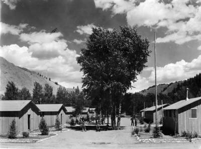 A group of CCC men stand in the clearing between two rows of barracks at a CCC Camp. In the middle of the clearing there are a few trees. Around each of the barracks is some landscaping, small trees and borders of white rocks.