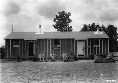 CCC officers posed with a dog and seated in front of the officer barracks. Sign in the center reads: 'Tepee of Big Chiefs' while the sign to the right of the photo reads: 'U.S. Forest Service'.