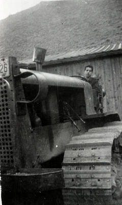 A CCC man sits in the driver's seat of a bulldozer in front of a building and hill.