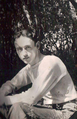 Close-up photo of a CCC man. He is wearing a long-sleeved white shirt and khakis. He is also wearing a necklace. Photo is taken in front of a thicket or bush.