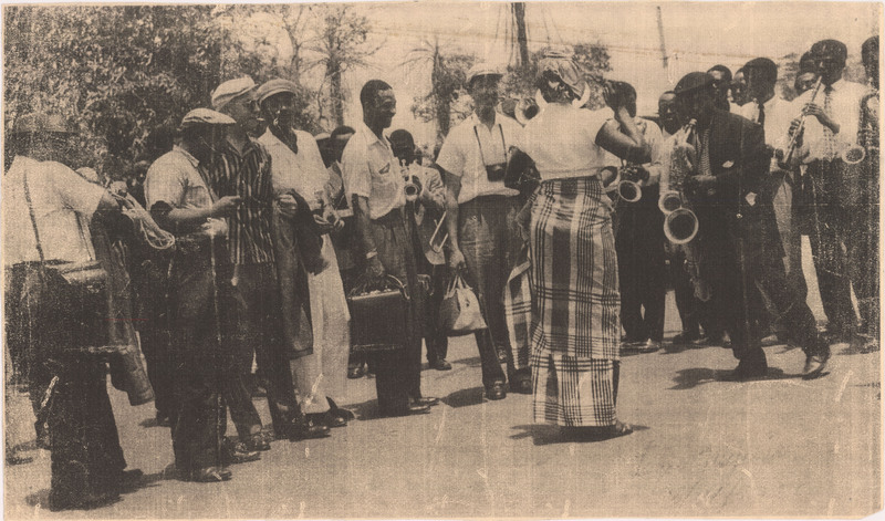 Doc Cheatham (center), a camera around his neck, smiles with Wilbur de Paris's (1900-1973) band on tour in Lagos, Nigeria. Several men stand outside in a crowd; a few are seen playing woodwind and brass instruments. In front of the men is a woman dancing, wearing a short-sleeved shirt, long skirt, and gele (head wrap). In the bottom right corner are traces of pencil marks; a clearer scan in Cheatham's I Guess I'll Get the Papers and Go Home, reveals that this says "Lagos, Nigeria  3/14/37."