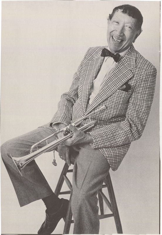 Doc Cheatham smiles while sitting on a wooden stool. He is angled toward the camera. Cheatham has dark curly hair, and wears a dress shirt, plaid blazer with pocket square, striped bowtie, slacks, socks, and dress shoes. His left hand, holding his trumpet, is crossed over his left hand, on which his wedding ring is visible. A Louis Armstrong pendant hangs from the trumpet. Photographer Scott Sternbach wrote "I knew him pretty well as I was moonlighting as a bartender at Sweet Basil Jazz Club...It was a 4th flr walkup but that did not deter Doc on his 80th Birthday. I had invited him up to the studio one Sunday to create an image for a commemorative poster. If I recall we had a birthday cake and a small celebration in my studio. I really love that image and in particular the way the Louis Armstrong pendant [sic] hangs in just the right place off of the horn."