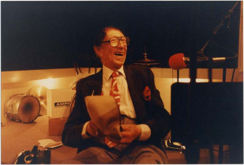Doc Cheatham lounges in a chair and laughs at something to the right of the camera. Cheatham wears a black jacket, khaki pants, white dress shirt, red tie with trumpets, drums, double basses, and piano keyboards on it, and has a red pocket-square. In front of him is a microphone covered in a red windscreen. Behind him to the right is an assembled open high-hat, and to the left is a silver tom drum with a worn head and a cymbal resting on the wall. 