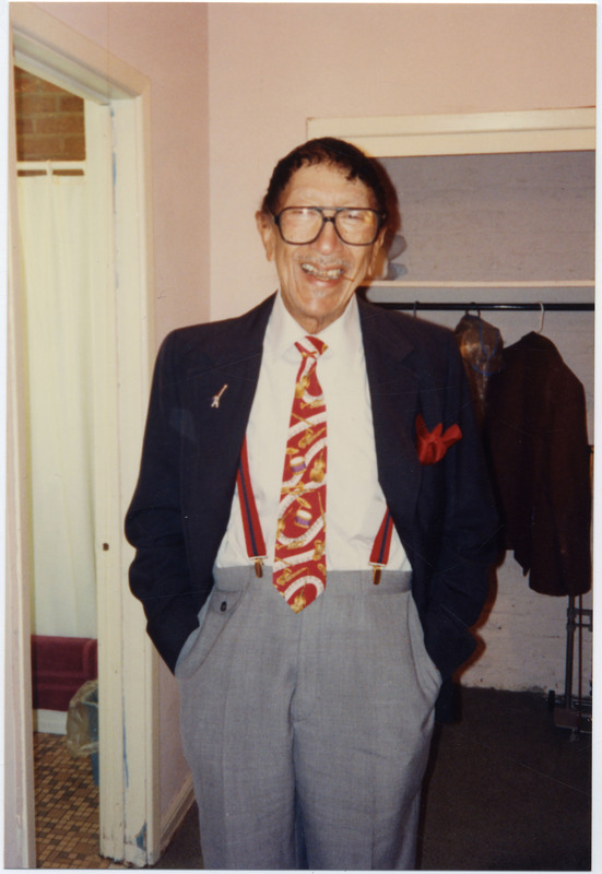 Doc Cheatham stands with both hands in pockets of his grey dress pants. He smiles with a toothpick between his teeth. He also wears black-rimmed glasses, red suspenders with a vertical navy stripe down the middle and gold clips, a red tie with various musical instruments on it, a navy jacket, and has a red pocket-square in his left pocket and a pin on the right side. Cheatham stands in front of a closet with coats hanging on a rack, and to the left, the tiled floor, bach mat, shower curtain, and trash can of a bathroom is visible.