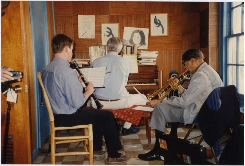 On the left, Brian O'Connell plays clarinet while sitting on a wooden chair with his back to the camera. On the right, Doc Cheatham plays trumpet with his right hand while holding and looking at sheet music in his left hand. He sits in a folding chair with a black vest and blue sweater draped over it. Cheatham's hair is combed from the back to the front of his head. He wears grey dress clothes. Between O'Connell and Cheatham is pianist Butch Thompson (1943-2022), playing on a wooden upright piano. On sticky note on a different picture of this room is written, "Brian and Hiroko's piano room," presumably that of O'Connell and his wife, Hiroko.
