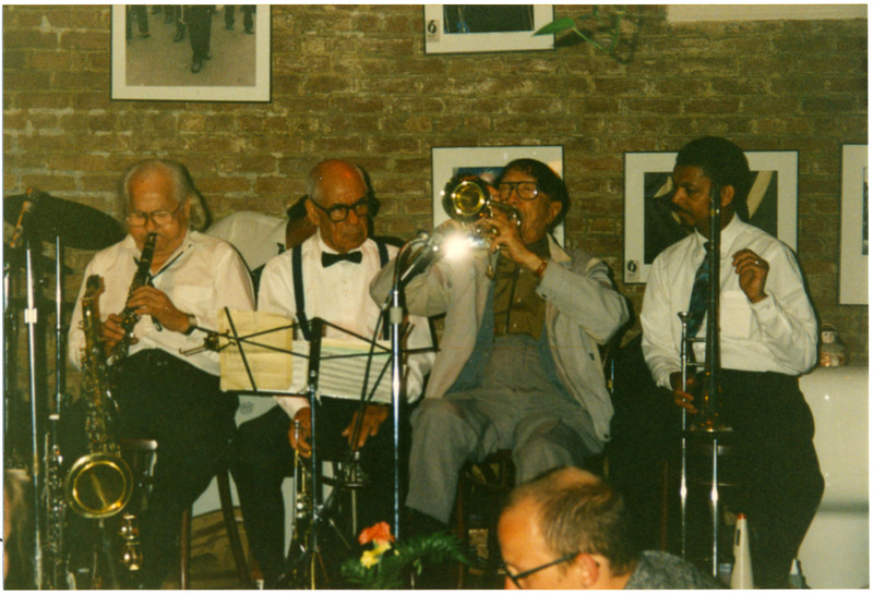 Doc Cheatham plays his trumpet aimed forward, parallel to the ground. Second from the right, Cheatham is sitting on a wooden stool and wears a brown dress shirt, black bolo tie, brown watch, grey jacket and pants, and black socks and shoes. To the left are Pud Brown (1917-1996) and trumpeter Lionel Ferbos (1911-2019), wearing white dress shirts, black ties, and pants. Behind and between them is Ernie Ellie (1942-) behind a drumset. The almost-bald Lionel Ferbos holds his trumpet down and watches Doc Cheatham seriously through black-rimmed glasses.  On the far left, Brown plays clarinet intently, eyes closed behind wire-frame glasses. In front of him are a saxophone and clarinet on stands. To the right of Cheatham is trombonist Fred Lonzo (1950-) pointing his instrument down and watching Cheatham. Behind the musicians is a brick wall with large framed photographs in the Palm Court Jazz Cafe.