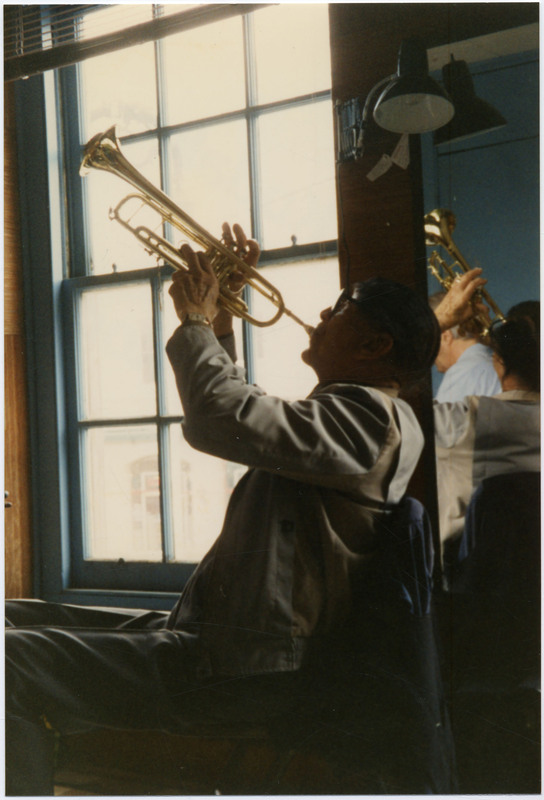 Doc Cheatham aims his trumpet up to the heavens in a practice session. In the mirror to the right of him, pianist Butch Thompson (1943-2022) can be seen. Behind Cheatham's left is a big, six-pane window with a blue wooden frame. Clarinetist Brian O'Connell is also present.