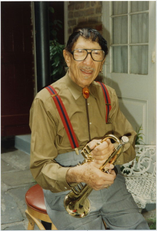 Doc Cheatham sits on a wooden stool with brown leather top, holding his trumpet in both hands. He wears black-rimmed glasses, a brown bolo tie, lighter brown dress shirt, red suspenders with a navy stripe down the middle and gold clips, and grey dress pants. Cheatham is outside a brick building in the daylight.