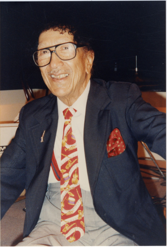 Doc Cheatham sits in a chair and smiles at a recording session. Cheatham wears black-rimmed glasses, a white dress shirt, red tie with various musical instruments, navy jacket with red pocket-square, grey pants, and black socks and shoes.