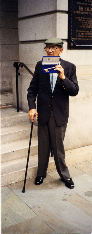 Doc Cheatham wears an olive green cap, black-rimmed glasses, bolo tie, light blue dress shirt, black suit jacket, dark grey slacks, black shiny dress shoes, and holds a black cane in his right hand. He stands outside of the Savannah Old City Hall in Savannah, GA and looks to the right. In his left hand is a Key to the City of Savannah on blue velvet backing. Cheatham received the key in 1996 with fellow musicians James Moody (1925-2010), Jon Faddis (1953-), and Irene Reid (1930-2008). Behind Cheatham are stone steps with a black railing and a stone pillar. On the pillar is the Savannah Historical Marker, commemorating the first steamship to cross the ocean.
