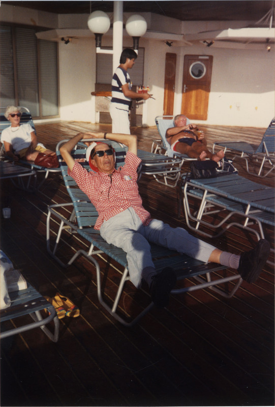 Doc Cheatham lounges in a pool chair, stretching out his legs. He wears a hat, red and white checkered short sleeve button-up, bolo tie, blue and white striped trousers, grey socks, black shoes, and sunglasses. He was a performer on this eighth annual Floating Jazz Festival run by the Norwegian Cruise Line.