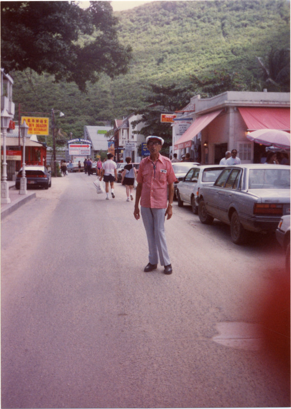 Doc Cheatham poses for a picture on a street in Philipsburg, capital of Sint Maarten. He wears a hat, red and white checkered short sleeve button-up, bolo tie, blue and white striped trousers, gold watch, black shoes, and sunglasses. In his left breast pocket is a photo or piece of paper. To the left and behind him is Sr. Marie Laurence Primary School, as well as a Golden Dragon Chinese Restaurant. In the background is a sign for the popular tourist destination Bobby's Marina, and in the distance is a tree-covered hill. To the right, lining the street, are parked cars. Sint Maarten was a stop on this November 1990 Norwegian Cruise Line journey to the Bahamas.