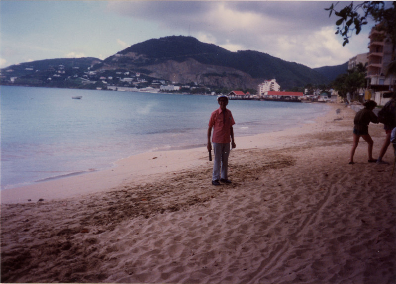 Doc Cheatham stands on the Great Bay Beach in Sint Maarten. He wears a hat, red and white checkered short sleeve button-up, bolo tie, blue and white striped trousers, gold watch, black shoes, and sunglasses. In his left breast pocket is a photo or piece of paper. In the background is water, buildings lining the beach, and foliage-covered hills. Sint Maarten was a stop on this November 1990 Norwegian Cruise Line journey to the Bahamas.