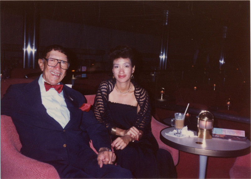 Doc Cheatham and his daughter, Alice, smile as they sit aboard red chairs on a cruise ship. He wears black-rimmed glasses, a navy suit jacket and pants, a white button-up, a gold watch, a red bowtie with polka dots, and a red pocket-square. On his right, Alice wears gold jewelry, a black lace shawl, and a black long dress with spaghetti straps. On the circular table to the left of them is a glass partially filled with liquid, and a menu. Alice Cheatham Croker and Amanda "Nellie" Cheatham (Croker's mother and Doc's widow), accepted the Grammy Doc Cheatham received posthumously in 1998.