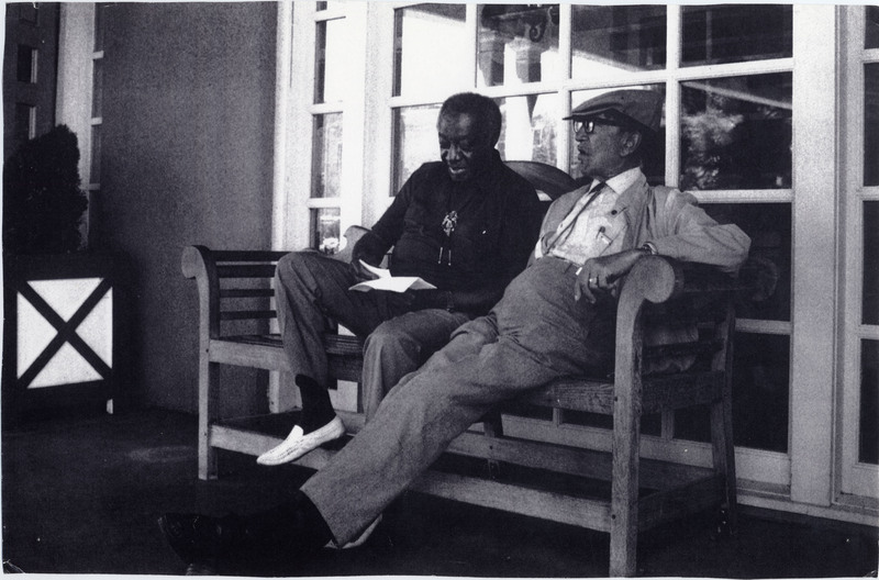Doc Cheatham (right) and Milt Hinton (left) sit on a wooden bench outside of a building with lots of paned windows. They are both wearing dress shirts, slacks, bolo ties, and formal shoes. Cheatham wears a watch, a cap, and rimmed glasses. He appears to be in conversation with Hinton, who is looking down at papers he holds in his hands. The picture has four small holes poked through the corners, such as those made by thumbtacks.