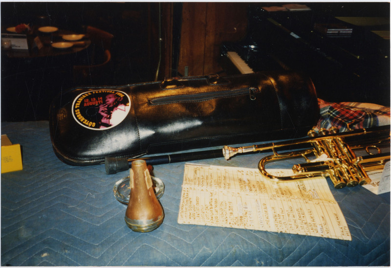 Photo of Doc Cheatham's possessions on a blue, quilt-covered table. There is a black, closed trumpet case with a sticker from the Göteborg Tradjazz Festival 1994, a trumpet with attached mouthpiece, trumpet mute (given to Cheatham by "King" Oliver), black cane, glass ashtray, music manuscript book index, and a plaid-patterned fabric on a blue, quilt-covered table. In the background on the right side, is an upright piano.