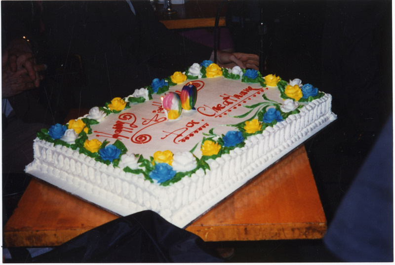 Photo of Doc Cheatham's 90th birthday cake. The cake is decorated with white piping around the outside edges, the top has alternating yellow, blue, and white flowers, green leaves, multicolored lit 90 candles on top, and in red is written, in cursive, "Happy Birthday Doc Cheatham" with lines and dots under his name. The cake is angled on a wooden table. 