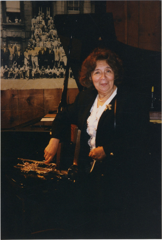 Photo of Doc Cheatham's wife, Amanda "Nellie" Cheatham smiling next to the table touching Doc's trumpet. She wears a single-strand pearl necklace, white buttoned shirt with lace down the front, a black formal jacket, and a gold pin with three flowers on her left side. In the background is a black grand piano with the top open. Behind the piano on the wooden wall is a copy of the 1958 "A Great Day in Harlem" black-and-white photograph, taken with 57 jazz musicians by Art Kane.