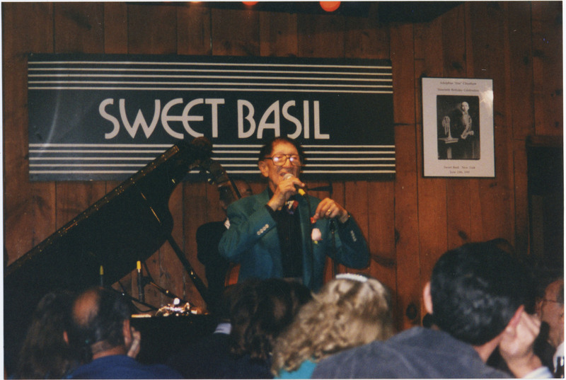 Doc Cheatham holds a microphone in his right hand as he speaks into it and holds a pink charm in his left hand. He wears black-rimmed glasses, a multi-colored bowtie, a dark blue suit jacket with red pocket square, a black button-down dress shirt, and a trumpet pin. In front of him, the backs of the heads of attendees can be seen. Behind Cheatham to the left is a black grand piano with the top open. On the wood-panelled wall behind is a black and white Sweet Basil sign, and to the left is a framed black and white poster that says "Adolphus 'Doc' Cheatham Ninetieth Birthday Celebration," a picture of Cheatham in a suit holding a saxophone with a coronet on a stand next to him taken at Lincoln Elementary, Nashville, Tennessee, and "Sweet Basil, New York June 13th, 1995."