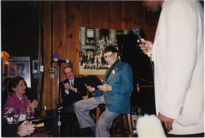 Doc Cheatham sits on a chair and smiles at a black rectangular object he holds in his hands. He wears black-rimmed glasses, a multi-colored bowtie, a wedding ring, a dark blue suit jacket with red pocket square, a black button-down dress shirt, grey slacks, and a trumpet pin. A microphone on a stand with yellow tape wrapped around the end is in front of him. Behind him to the left, pianist Chuck Folds (1938-2022) smiles and claps while sitting at a black grand piano. He wears a checkered tie, button-down shirt, and black blazer. Behind the piano on a wood-panelled wall is the 1958 "A Great Day in Harlem" photograph by Art Kane. On the left, a framed Greenwich Village Jazz Festival is hung on a wall. In front are attendees with their hands clapping. On the right, an announcer in a white suit holds a microphone with a yellow tape-wrapped end.