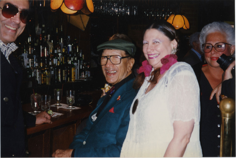 Doc Cheatham sits at a bar and smiles towards the camera. He wears black-rimmed glasses, a multi-colored bowtie, a dark blue suit jacket with red pocket square, a black button-down dress shirt, grey slacks, and a trumpet pin. On Cheatham's right is a middle-aged Caucasian woman with two pink ribbons in her brown hair, large shiny earrings, pink lipstick, lilac eyeshadow, and wears a white lace top with a gold pin in the top center. At the left edge is a man smiling at the camera. He wears brown sunglasses, a blue and white button-down, blue blazer, and his fist is closed on the wooden bar counter. 