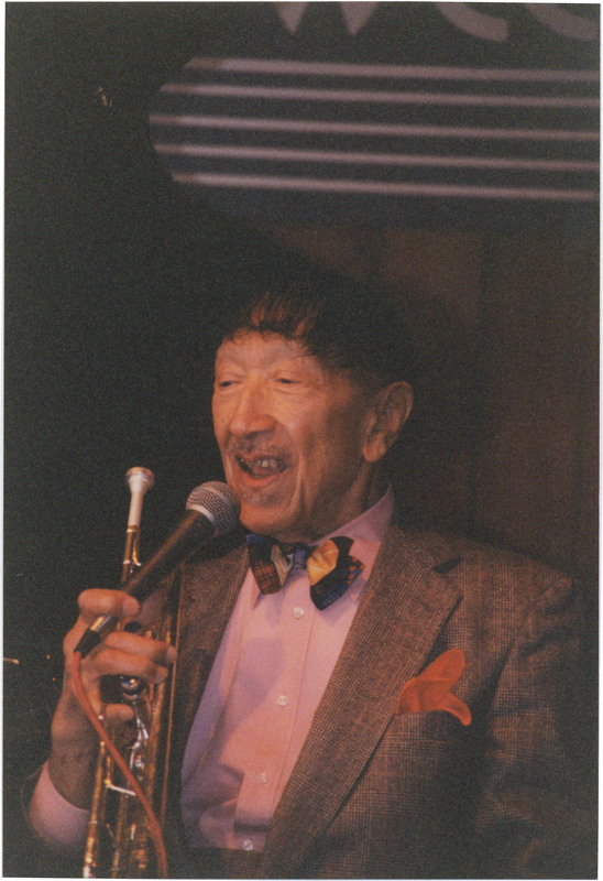 Doc Cheatham speaks into a microphone, which he holds with his left hand along with his trumpet. He wears a pale pink button-down, multicolored bow tie, plaid-patterned jacket, and red pocket-square. Behind him is a wood-panelled wall with a black and white Sweet Basil sign, and the top of an open black grand piano. Cheatham and his quartet played regularly at Sweet Basil Jazz Club at jazz brunches for 17 years. Sweet Basil was a prominent location for live jazz performances from when it opened in 1974 till it closed in April 2001.
