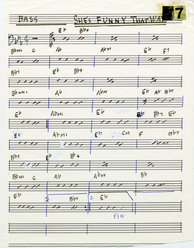 Loose page with printed music staves. At the top, "Bass" is written and underlined, and "She's funny that way" is written and underlined twice in black marker. In the top right corner are "5" and "7" gold reflective stickers. Written in black marker is a bass clef, rests, dashes to indicate eighth notes, repeat measure signs, repeat barlines, and chord names over measures. The key signature (Bb, Eb, Ab) and time signature (cut time) are written in black pen. Measure lines, the 1st and 2nd endings, and "FIN" at the end are written in blue pen ink. There are several white-out marks on the page. "She's funny that way" (1929) was composed by Charles N. Daniels (pseudonym: Neil Moret; 1878-1943) and Richard Whiting (1891-1938) for the short film "Gems of M-G-M." Cheatham released a recording of this piece on his record It's a Good Life (1982), and Cheatham and Nicholas Payton (1973-) released a recording on Doc Cheatham & Nicholas Payton (1997).