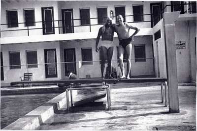 Doc Cheatham poses with Eddie Barefield (1909-1991) in swimming attire. The two performed in Cab Calloway (1907-1994)'s jazz band together and were there on tour along with Milt Hinton (1910-2000), his wife Mona Hinton (19192008), and others. Barefield was a saxophonist, clarinetist and arranger who also worked with Louis Armstrong (1901-1971), Ella Fitzgerald (1917-1966), and Duke Ellington (1899-1974). The musicians stand on a diving board extending over a pool. In the background, a person sits at the edge of the pool. Behind is part of the Sir John Hotel. To the right of Cheatham is a sign reading "Shower before entering pool." The Sir John Hotel, which opened in 1951 under the moniker Lord Calvert Hotel, hosted many famous people, such as Nat King Cole, Ella Fitzgerald, Cab Calloway, and Muhammed Ali until it was torn down in the 1960s. Milt Hinton, bassist, and photographer, wrote in his book Playing the Changes, "We were staying at a brand new black motel called the Sir John. It was considered luxurious in those days because it had a pool and its own little nightclub," (pg 136), the Knight Beat Club. The hotel was popular in Overtown, an area of segregated Miami where people of color were allowed to stay. The picture has four small holes poked through the corners, such as those made by thumbtacks.