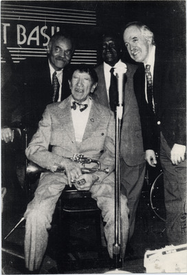 Doc Cheatham sits and holds his trumpet. From left to right, Cheatham is surrounded by Jackie Williams (1933-), Bucky Calabrese (1927-1995), and Chuck Folds (1938-2022). They made up Doc's Quartet, performing on regular Sunday afternoons at the popular jazz restaurant Sweet Basil in Greenwich Village, New York. The right side edge has "Folds" written on it, presumably in reference to Jane Folds (-2022), the photographer and Chuck's wife. The picture has four small holes poked through the corners, such as those made by thumbtacks.