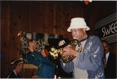 Doc Cheatham sits on a chair and plays his trumpet pointing towards the ceiling.  He wears black-rimmed glasses, a multi-colored bowtie, a dark blue suit jacket with red pocket square, a black button-down dress shirt, and a trumpet pin. A microphone on a stand with yellow tape wrapped around the end is in front of him. Behind him to the left, pianist Chuck Folds (1938-2022) plays on a black grand piano. Behind the piano on a wood-panelled wall is the 1958 "A Great Day in Harlem" photograph by Art Kane. Folds wears a checkered tie, button-down shirt, and black blazer. On the piano is a dark orange plunger mute. Another microphone is pointed towards the inside of the piano. To the left in front of Cheatham is Al Grey (1925-2000), wearing a striped shirt, blue jacket, and white bucket hat and playing a bass trumpet. Behind Grey is Earl May (1927-2008) playing a bass.