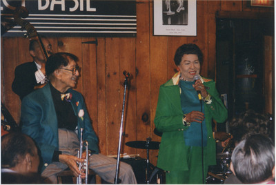 Doc Cheatham sits on a chair and smiles with his head turned to the left. He wears black-rimmed glasses, a multi-colored bowtie, a dark blue suit jacket with red pocket square, a black button-down dress shirt, grey slacks, and a trumpet pin. He holds his trumpet pointed towards the floor with both hands. Cheatham looks at a woman with short black hair, red flower earrings, a teal sweater, green suit jacket and pants, a beaded necklace, red lipstick, and a chunky silver ring, who speaks into a microphone she holds with her left hand. Behind her is a drum set and drummer Jackie Williams (1933-) 's hands can be seen. Behind Cheatham on the left is Earl May (1927-2008) holding his bass. On the wood-panelled wall behind everyone is a black and white Sweet Basil sign, and a framed poster to the left of it with a photo of a young Cheatham with a saxophone and cornet. The backs of attendee's heads can be seen in the foreground.