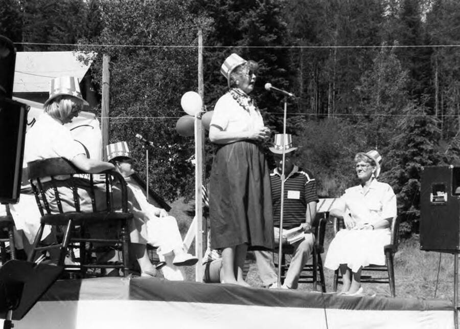 A woman speaking on a stage during Bovill's Statehood Day celebration, while others sit behind her.
