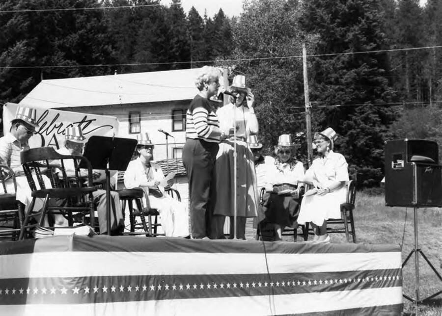 Two women speaking on a stage while others sit behind them.
