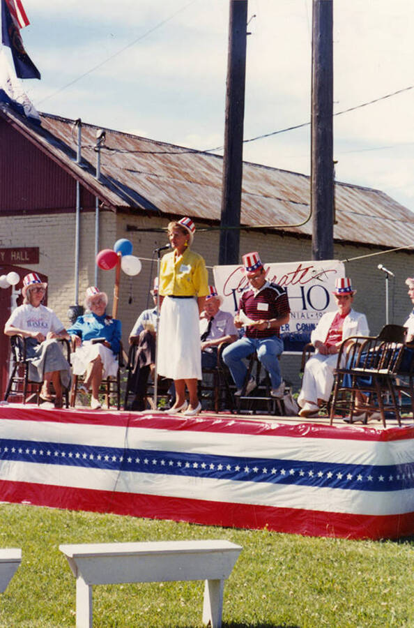 Carol Reynolds speaks on a stage during Bovill's Statehood Day celebration, while others sit in the backround of the stage.