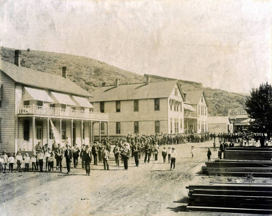 Crowd of people in street with band in foreground. DeLamar Mercantile in background. DeLamar, Idaho.